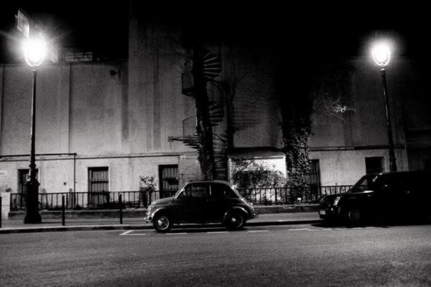 A small dark car parked on an empty night street between two bright lamps, with a spiral stair on the building behind.